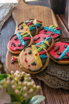 Colorful cookies decorated with icing to resemble faces with distinctive features, such as red lips, black eyes, and multicolored headdresses. The cookies are placed on a wooden cutting board, with intricate doily details beside them, creating a playful and artistic presentation.