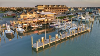 A marina with wooden docks and numerous boats moored along the calm water. A large, multi-storied building, possibly a restaurant or marina office, is visible in the background with an American flag hanging in front. The late afternoon sunlight casts a warm glow over the scene, highlighting the boats and the reflections in the water.