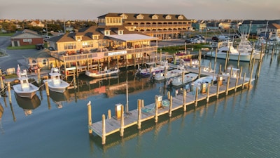 A marina with wooden docks and numerous boats moored along the calm water. A large, multi-storied building, possibly a restaurant or marina office, is visible in the background with an American flag hanging in front. The late afternoon sunlight casts a warm glow over the scene, highlighting the boats and the reflections in the water.