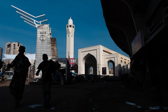 An urban scene featuring a large architectural gateway structure with an elegant design. To the right, a tall white clock tower stands prominently against the clear blue sky. The foreground includes silhouettes of people walking, adding a sense of scale and movement to the scene.