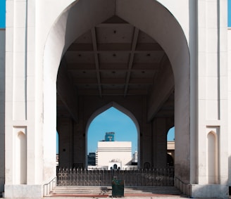 An architectural structure with large arched openings, constructed from light-colored materials. The design includes intricate geometric patterns and symmetrical lines. In the background, a modern urban landscape featuring several buildings.