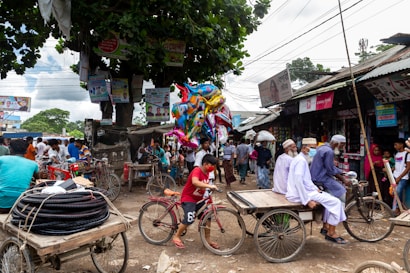 A bustling street market scene with numerous people engaged in various activities. Stalls with goods line the street, and vendors interact with customers. Colorful balloons are carried by a vendor, and bicycles are a common mode of transport. A group of men wearing traditional clothing sit on a rickshaw, while others walk about or stand near shops.