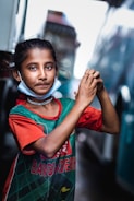 A young girl stands outdoors, wearing a green and red jersey with 'Bangladesh' written on it. She has a relaxed expression, with her hands clasped together in front of her body. The girl also has a mask around her neck and a simple necklace. The background shows blurred city structures and possibly a vehicle.