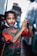 A young girl stands outdoors, wearing a green and red jersey with 'Bangladesh' written on it. She has a relaxed expression, with her hands clasped together in front of her body. The girl also has a mask around her neck and a simple necklace. The background shows blurred city structures and possibly a vehicle.