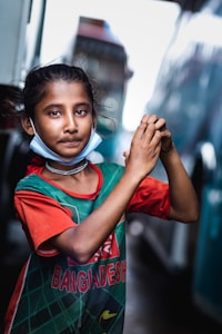 A young girl stands outdoors, wearing a green and red jersey with 'Bangladesh' written on it. She has a relaxed expression, with her hands clasped together in front of her body. The girl also has a mask around her neck and a simple necklace. The background shows blurred city structures and possibly a vehicle.
