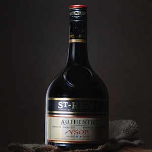 A bottle of St-Rémy Authentic French Brandy is set against a dark background. The bottle is elegantly designed with a black label featuring gold and red accents. The lighting highlights the curves and label details of the bottle, creating a sophisticated and classic appearance.