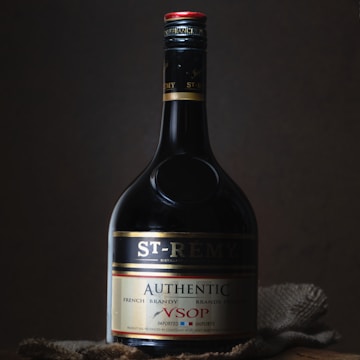 A bottle of St-Rémy Authentic French Brandy is set against a dark background. The bottle is elegantly designed with a black label featuring gold and red accents. The lighting highlights the curves and label details of the bottle, creating a sophisticated and classic appearance.