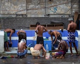 A group of men are gathered around an outdoor bathing area, engaging in activities such as washing and rinsing under a makeshift tap. The setting suggests a communal bathing space with a worn, urban backdrop. The men are using various items such as buckets and containers to assist in their washing.