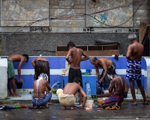 A group of men are gathered around an outdoor bathing area, engaging in activities such as washing and rinsing under a makeshift tap. The setting suggests a communal bathing space with a worn, urban backdrop. The men are using various items such as buckets and containers to assist in their washing.