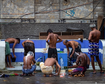 A group of men are gathered around an outdoor bathing area, engaging in activities such as washing and rinsing under a makeshift tap. The setting suggests a communal bathing space with a worn, urban backdrop. The men are using various items such as buckets and containers to assist in their washing.