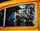 A person cleans the interior of a yellow taxi, with reflections of trees visible on the car window. The person appears to be focused on their task, using a red cloth to wipe the inside of the window. There is a blue car parked in the background.