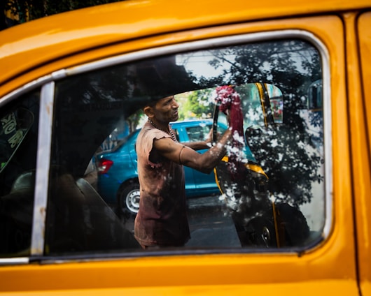 A person cleans the interior of a yellow taxi, with reflections of trees visible on the car window. The person appears to be focused on their task, using a red cloth to wipe the inside of the window. There is a blue car parked in the background.