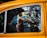 A person cleans the interior of a yellow taxi, with reflections of trees visible on the car window. The person appears to be focused on their task, using a red cloth to wipe the inside of the window. There is a blue car parked in the background.