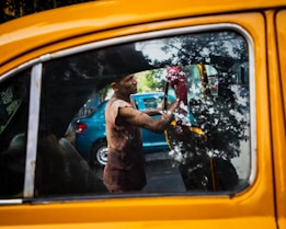 A person cleans the interior of a yellow taxi, with reflections of trees visible on the car window. The person appears to be focused on their task, using a red cloth to wipe the inside of the window. There is a blue car parked in the background.