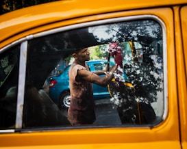 A person cleans the interior of a yellow taxi, with reflections of trees visible on the car window. The person appears to be focused on their task, using a red cloth to wipe the inside of the window. There is a blue car parked in the background.