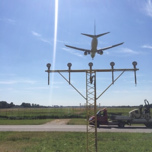 An airplane is captured flying low over an approach light system at an airport. The sky is clear with a few wispy clouds, and a road and grassy field are visible below. Two vehicles are parked nearby, including a red van and a utility truck.