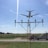 An airplane is captured flying low over an approach light system at an airport. The sky is clear with a few wispy clouds, and a road and grassy field are visible below. Two vehicles are parked nearby, including a red van and a utility truck.
