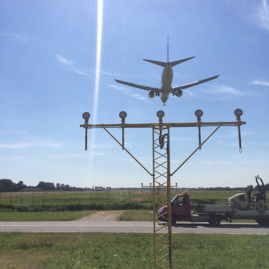 An airplane is captured flying low over an approach light system at an airport. The sky is clear with a few wispy clouds, and a road and grassy field are visible below. Two vehicles are parked nearby, including a red van and a utility truck.