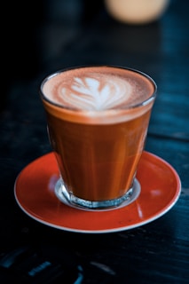 A glass of coffee with latte art on top is placed on an orange saucer. The coffee has a creamy texture with a white, intricate leaf pattern created using steamed milk. The glass and saucer are set against a dark, blurred background, emphasizing the warm tones of the coffee.