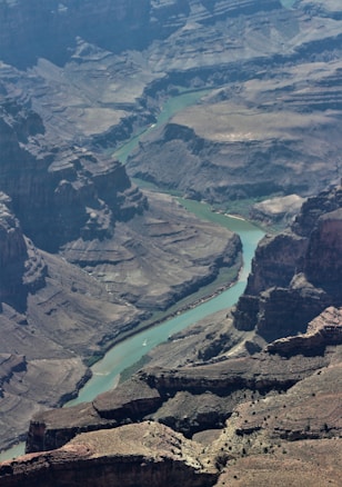 A vast, rocky canyon stretches out, carved by a winding river that snakes its way through the landscape. The river appears calm and meanders between steep, rugged cliffs and layered rock formations. The geological formations display a variety of textures and colors, creating a dramatic and expansive view.