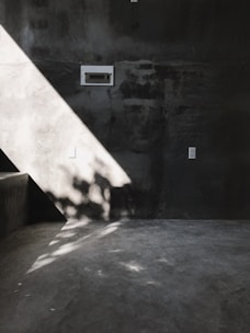 A minimalist interior features a concrete wall and floor, accented by a dramatic diagonal shaft of sunlight creating sharp shadows and highlights. A small electrical panel is mounted on the wall, with its subdued presence contributing to the stark, industrial aesthetic. The play of light and shadow adds depth to the otherwise muted setting.