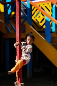 A child wearing a colorful outfit is holding onto a red pole. The background features a vibrant setting with geometric patterns in blue, yellow, and orange on a wooden structure.
