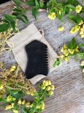A black, ergonomically shaped comb lies on a small burlap drawstring pouch. The setup is on a wooden surface, bordered by various green leaves and yellow flowers, creating a natural and rustic aesthetic.