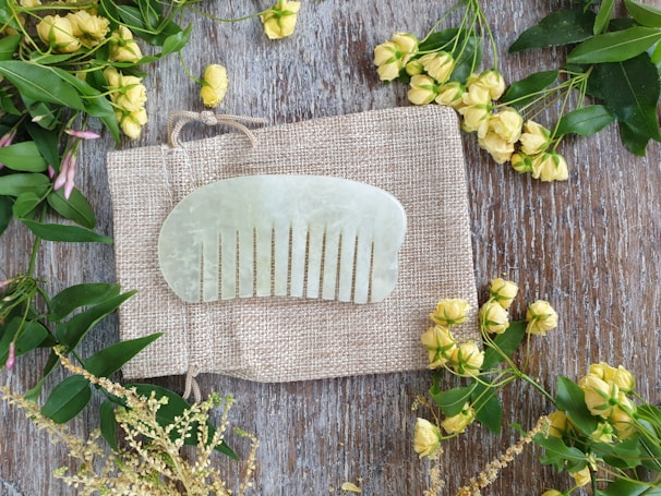 A jade comb is placed on a textured, woven fabric, surrounded by yellow flowers and green leaves on a wooden surface.