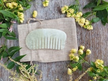 A jade comb is placed on a textured, woven fabric, surrounded by yellow flowers and green leaves on a wooden surface.