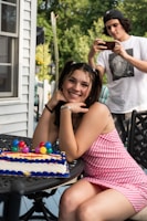 A young woman is sitting outdoors with a colorful birthday cake on a table. She is smiling and looking towards the camera, wearing a pink and white striped dress. In the background, a young person in a white t-shirt and cap is capturing a photo with a smartphone. The setting appears to be a backyard with green foliage and part of a house visible.