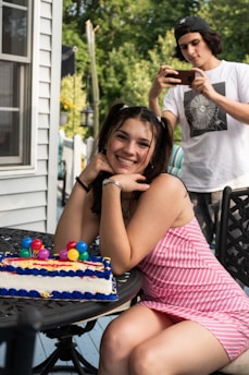 A young woman is sitting outdoors with a colorful birthday cake on a table. She is smiling and looking towards the camera, wearing a pink and white striped dress. In the background, a young person in a white t-shirt and cap is capturing a photo with a smartphone. The setting appears to be a backyard with green foliage and part of a house visible.