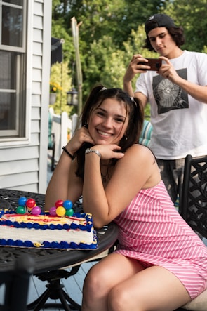 A young woman is sitting outdoors with a colorful birthday cake on a table. She is smiling and looking towards the camera, wearing a pink and white striped dress. In the background, a young person in a white t-shirt and cap is capturing a photo with a smartphone. The setting appears to be a backyard with green foliage and part of a house visible.