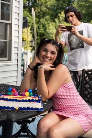 A young woman is sitting outdoors with a colorful birthday cake on a table. She is smiling and looking towards the camera, wearing a pink and white striped dress. In the background, a young person in a white t-shirt and cap is capturing a photo with a smartphone. The setting appears to be a backyard with green foliage and part of a house visible.