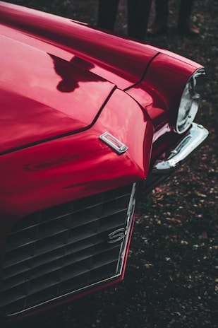 Close-up view of a vintage red car with a shiny finish. The image shows a detailed look at the front grille, a headlight, and part of the hood. The scene is set outdoors with a shadow visible on the car's surface, adding an artistic element.