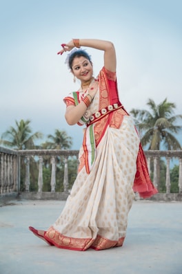 A woman dressed in a traditional Indian saree with intricate patterns dances gracefully on a terrace surrounded by palm trees. Her attire is richly adorned with a red and gold color scheme, and she wears traditional jewelry. The background shows a serene, clear sky.