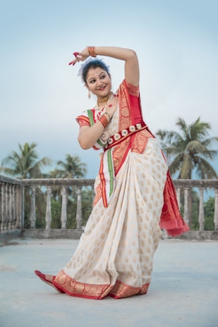 A woman dressed in a traditional Indian saree with intricate patterns dances gracefully on a terrace surrounded by palm trees. Her attire is richly adorned with a red and gold color scheme, and she wears traditional jewelry. The background shows a serene, clear sky.