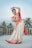 A woman dressed in a traditional Indian saree with intricate patterns dances gracefully on a terrace surrounded by palm trees. Her attire is richly adorned with a red and gold color scheme, and she wears traditional jewelry. The background shows a serene, clear sky.