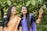 Two women dressed in traditional sarees are standing amidst lush green leaves. One woman is wearing an orange saree with floral patterns, and the other is wearing a purple saree. They appear to be smiling and looking in the same direction, with one of them pointing towards something.