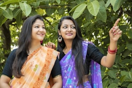 Two women dressed in traditional sarees are standing amidst lush green leaves. One woman is wearing an orange saree with floral patterns, and the other is wearing a purple saree. They appear to be smiling and looking in the same direction, with one of them pointing towards something.