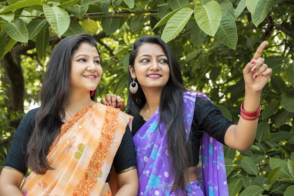 Two women dressed in traditional sarees are standing amidst lush green leaves. One woman is wearing an orange saree with floral patterns, and the other is wearing a purple saree. They appear to be smiling and looking in the same direction, with one of them pointing towards something.