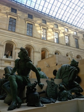 Large bronze sculptures of classical figures are set against a background of grand architecture with tall windows and a glass ceiling, casting shadows and illuminating the space.