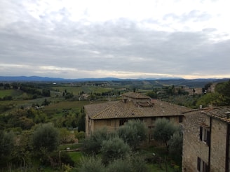 A picturesque landscape featuring a rustic stone house with a tiled roof in the foreground, surrounded by lush green trees and olive groves. The background displays rolling hills and fields extending into the distance under a cloudy sky.