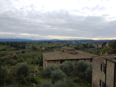 A picturesque landscape featuring a rustic stone house with a tiled roof in the foreground, surrounded by lush green trees and olive groves. The background displays rolling hills and fields extending into the distance under a cloudy sky.