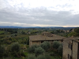 A picturesque landscape featuring a rustic stone house with a tiled roof in the foreground, surrounded by lush green trees and olive groves. The background displays rolling hills and fields extending into the distance under a cloudy sky.