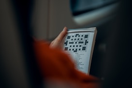 A person holds a book featuring a crossword puzzle with clues listed below the grid. The person is dressed in orange, and the focus is on the hands and puzzle.