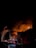 Firefighters are battling a large building fire at night. Tall plumes of smoke and flames engulf the structure as fire trucks, equipped with ladders and hoses, line the street. The scene is illuminated by flashing emergency lights and the glow of the fire.