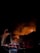 Firefighters are battling a large building fire at night. Tall plumes of smoke and flames engulf the structure as fire trucks, equipped with ladders and hoses, line the street. The scene is illuminated by flashing emergency lights and the glow of the fire.