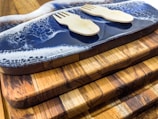 A set of wooden cutting boards stacked on top of each other with unique grain patterns. On the top board, there is a decorative resin wave design in blue and white. Two small wooden sporks are placed on the resin surface.
