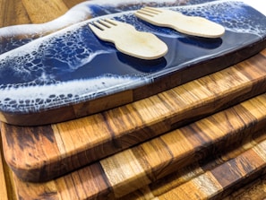 A set of wooden cutting boards stacked on top of each other with unique grain patterns. On the top board, there is a decorative resin wave design in blue and white. Two small wooden sporks are placed on the resin surface.