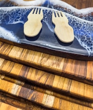 Two wooden sporks are placed on a dark resin cutting board with a wave-like design. The cutting board rests on several stacked wooden boards with varying shades of brown.